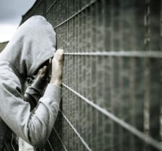 Photo of a person in a hoodie with their hands and head pressed against a metal fence.