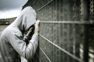 Photo of a person in a hoodie with their hands and head pressed against a metal fence.
