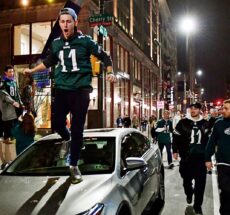 Photo of large crowd of NFL fans in the streets after a football game, one of whom is standing on top of a car.