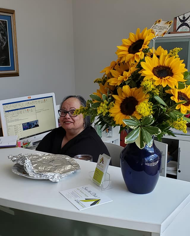 A smiling woman sitting at a desk by a computer with a vase of sunflowers.