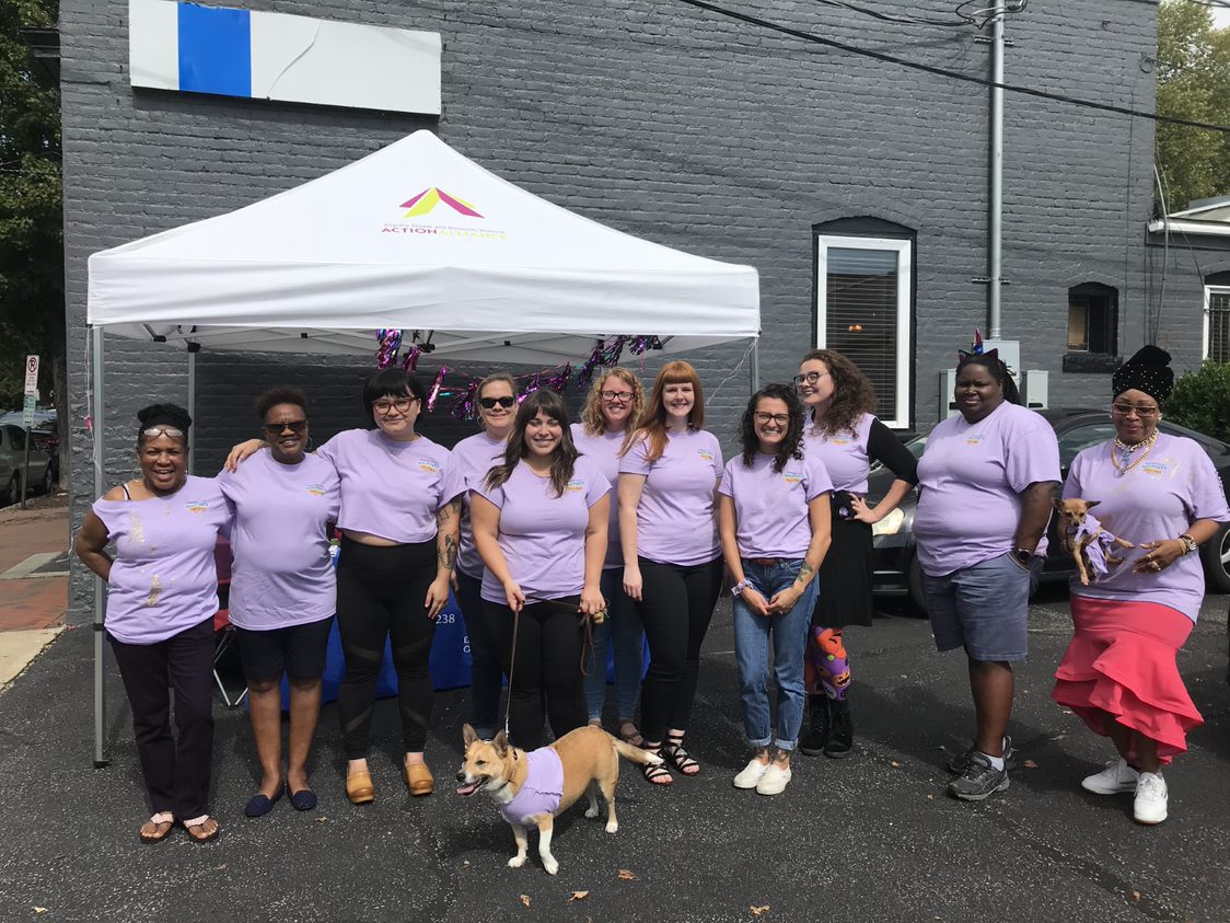 Group of 11 people and two dogs wearing purple EmPower Challenge t-shirts in front an Action Alliance tent.