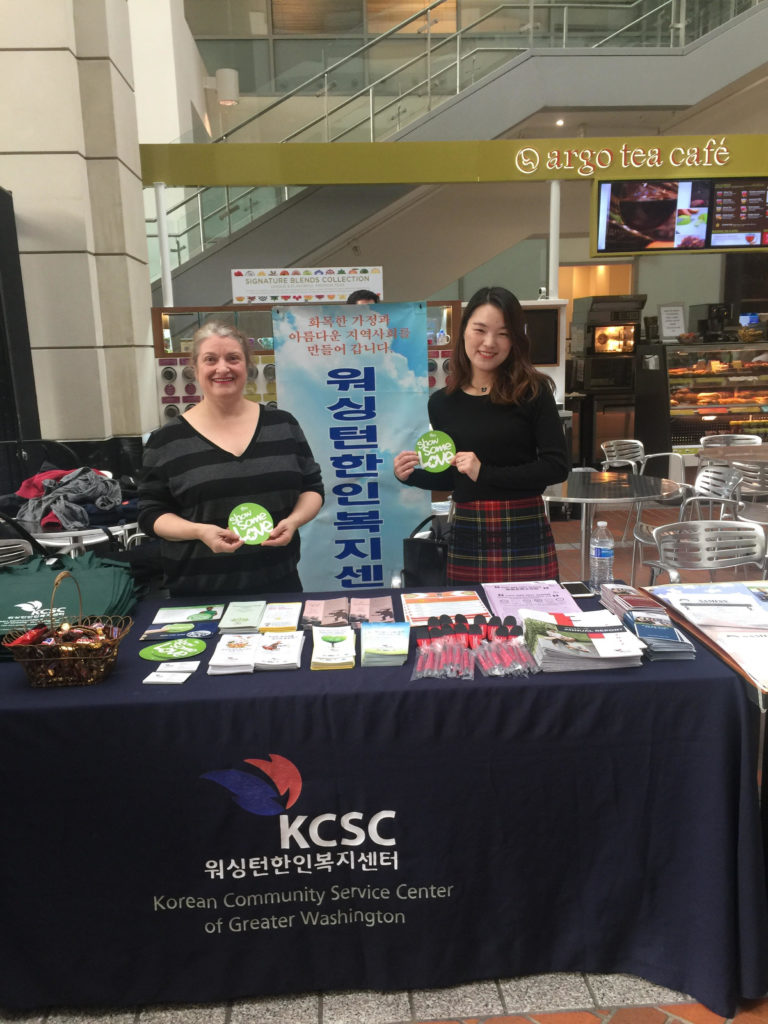 Two people standing behind a table with a dark colored tablecloth that has "KCSC- Korean Community Service Center of Greater Washington" on it and is covered with brochures and pamphlets to promote KCSC's services and information.