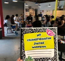 A hand holding a hand-drawn composition notebook with the title "My Transformative Justice Workbook" in the foreground, with a group of training participants in the background.