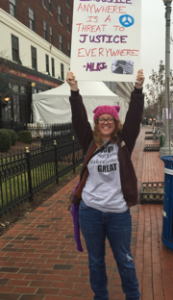 Photo of Katie Moffitt of the Action Alliance holding a sign that reads "Injustice anywhere is a threat to justice everywhere. -MLK Jr." and wearing a pink knit hat.