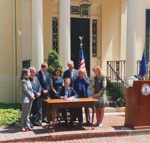 A group of seven men and women in suits and business attire surround Virginia's Gov. Northam as he signs into law HB 1992. Among the seven people is Del. Kathleen Murphy who cries tears of joy. The group is on a red brick sidewalk with the flags of the United States and Virginia in the background.